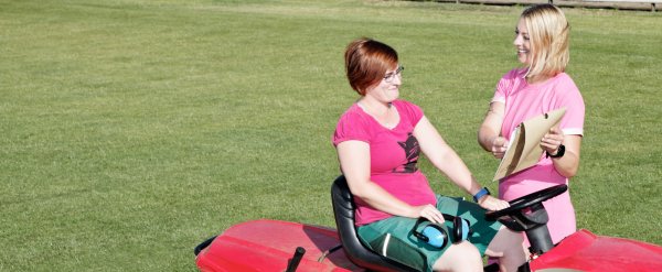 Teilnehmerin steht mit Aufsitzmäher auf Sportplatz und erhält Anleitung von Fachkraft.