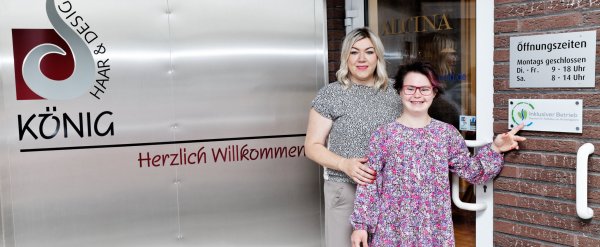 Teilnehmerin und Patin stehen neben dem Schild „Inklusiver Betrieb“ an einem Friseursalon.