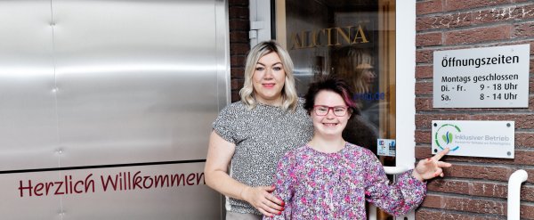 Teilnehmerin und Patin stehen neben dem Schild „Inklusiver Betrieb“ an einem Friseursalon.