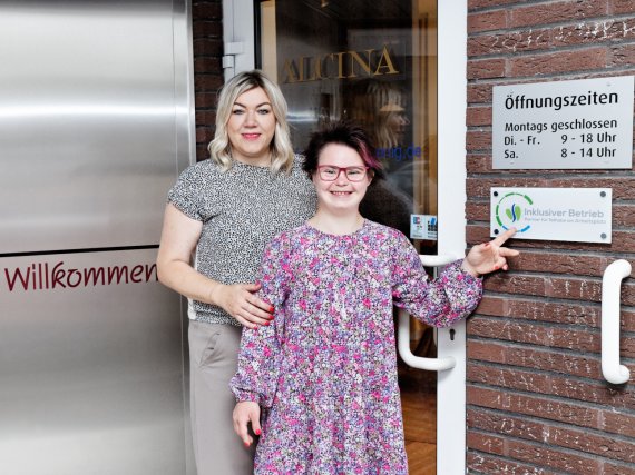 Teilnehmerin und Patin stehen neben dem Schild „Inklusiver Betrieb“ an einem Friseursalon.