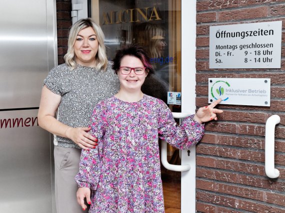 Teilnehmerin und Patin stehen neben dem Schild „Inklusiver Betrieb“ an einem Friseursalon.