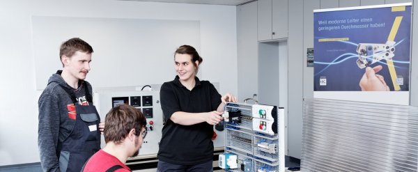 Ausbilderin erklärt zwei Auszubildenden einen Schaltkasten.