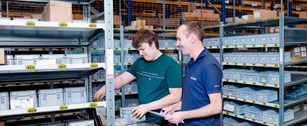 Vertreter des Betriebs und Auszubildender bei der Arbeit im Lager am Regal.