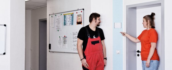Mitarbeiterin zeigt einem Auszubildenden auf einem Flur das Farbleitsystem.