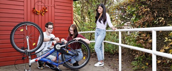 Zwei Jugendliche reparieren ein Fahrrad. Eine Mitarbeiterin schaut ihnen zu.