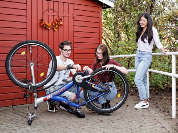 Zwei Jugendliche reparieren ein Fahrrad. Eine Mitarbeiterin schaut ihnen zu.