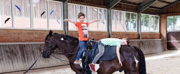 Zwei Kinder sitzen in einer Reithalle auf einem Pferd.