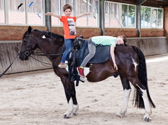 Zwei Kinder sitzen in einer Reithalle auf einem Pferd.
