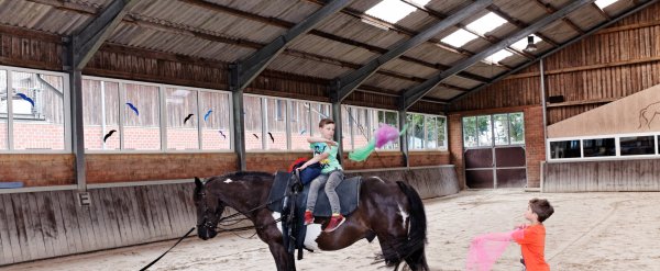 Zwei Kinder jonglieren in der Reithalle mit Tüchern. Eines sitzt auf einem Pferd.