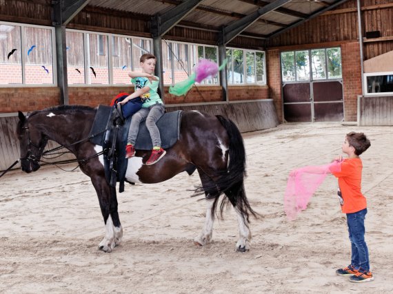 Zwei Kinder jonglieren in der Reithalle mit Tüchern. Eines sitzt auf einem Pferd.