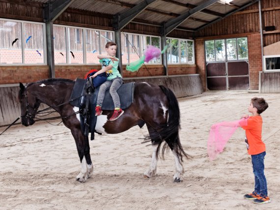 Zwei Kinder jonglieren in der Reithalle mit Tüchern. Eines sitzt auf einem Pferd.