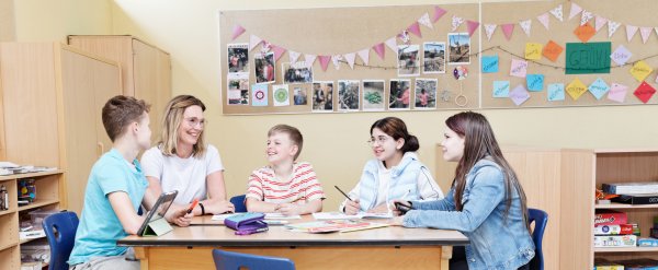 Mitarbeiterin sitzt mit vier Kindern in einem Klassenraum.