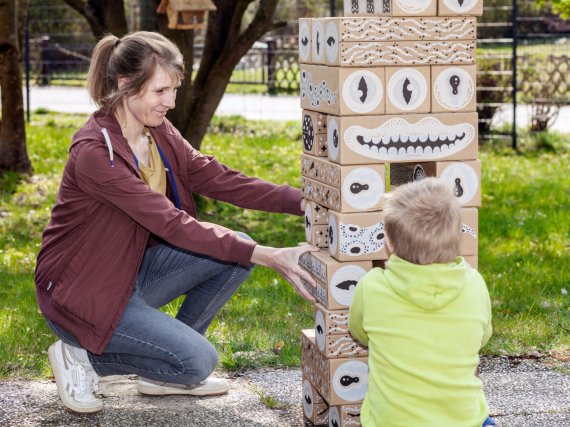 Mitarbeiterin und Kind spielen im Garten mit einem Turm aus großen Holzklötzen.