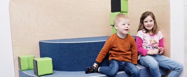 Zwei Kinder, eines mit Down-Syndrom, sitzen in der Kita nebeneinander auf einer Bank.