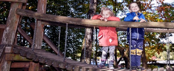 Zwei Kinder stehen auf dem Spielplatz auf einer Holzbrücke.