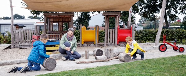 Zwei Kinder spielen auf einem Spielplatz mit großen Holzklötzen. Eine Erzieherin sieht ihnen zu.