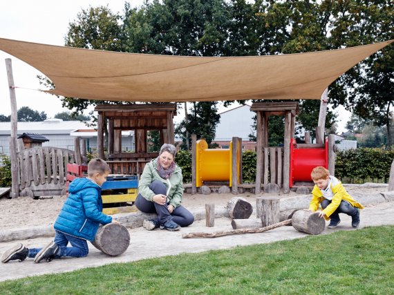 Zwei Kinder spielen auf einem Spielplatz mit großen Holzklötzen. Eine Erzieherin sieht ihnen zu.