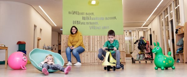 Zwei Kinder spielen im Flur eines Kindergartens. Eine Erzieherin schaut ihnen zu.