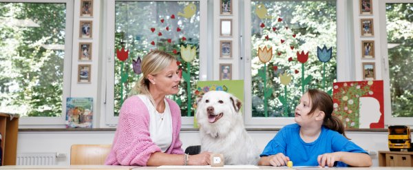 Lehrkraft und Schülerin spielen Lese-Lernspiel. Ein Hund sitzt zwischen ihnen.