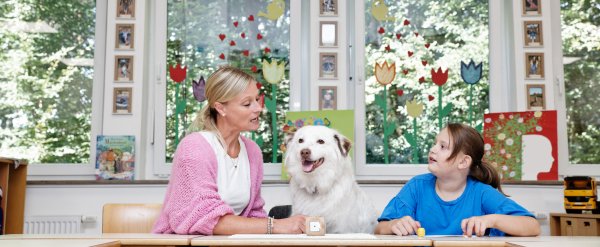 Lehrkraft und Schülerin spielen Lese-Lernspiel. Ein Hund sitzt zwischen ihnen.
