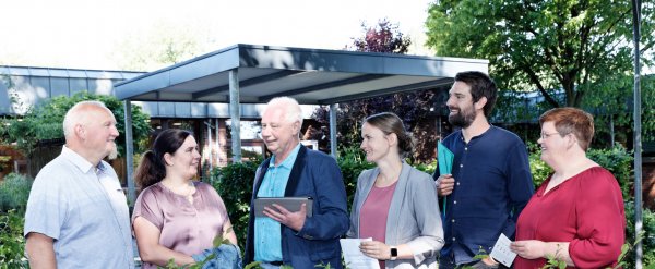 Sechs Personen stehen hinter einer niedrigen Hecke und schauen sich an.