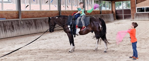 Zwei Kinder jonglieren in der Reithalle mit Tüchern. Eines sitzt auf einem Pferd.
