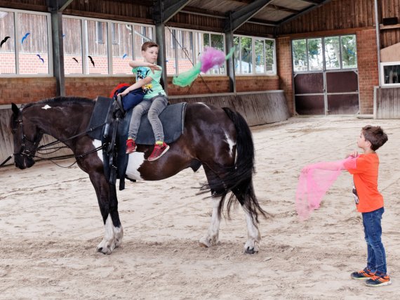 Zwei Kinder jonglieren in der Reithalle mit Tüchern. Eines sitzt auf einem Pferd.